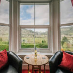 Bay window seating area in Room 5 at Bryn Bella Guest House, with red curtains and armchairs looking out over the wooded Conwy Valley.